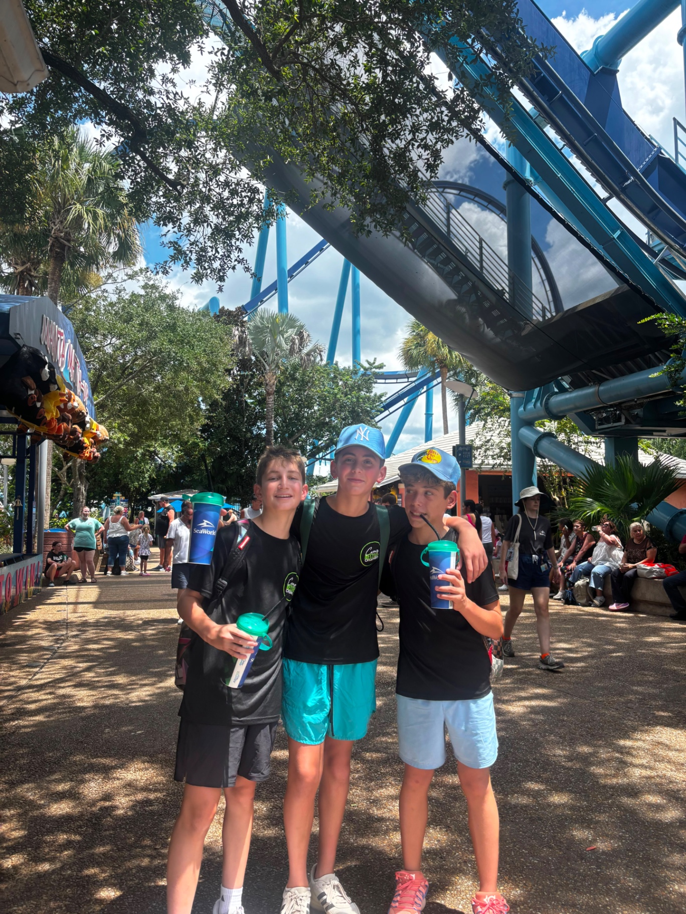 Three campers with souvenir cups beneath a blue roller coaster track