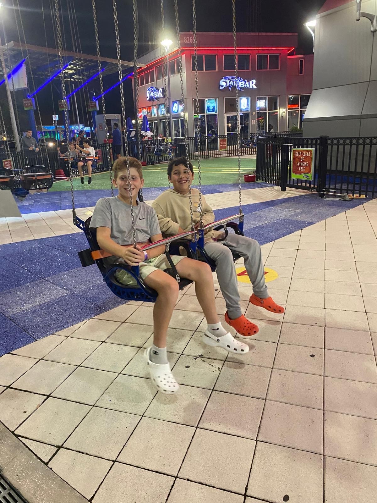 Two campers riding swing seats at night beside a lit sign