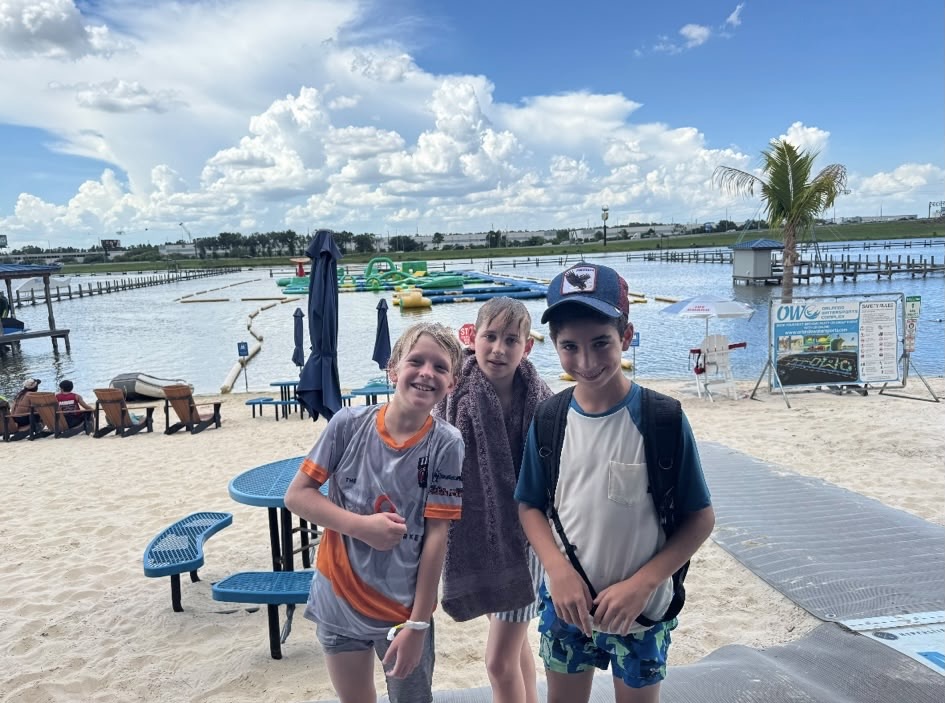 Three smiling campers on the beach in front of a lakefront waterpark