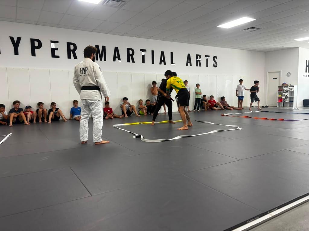 Two campers sparring on the mats while an instructor and seated students watch