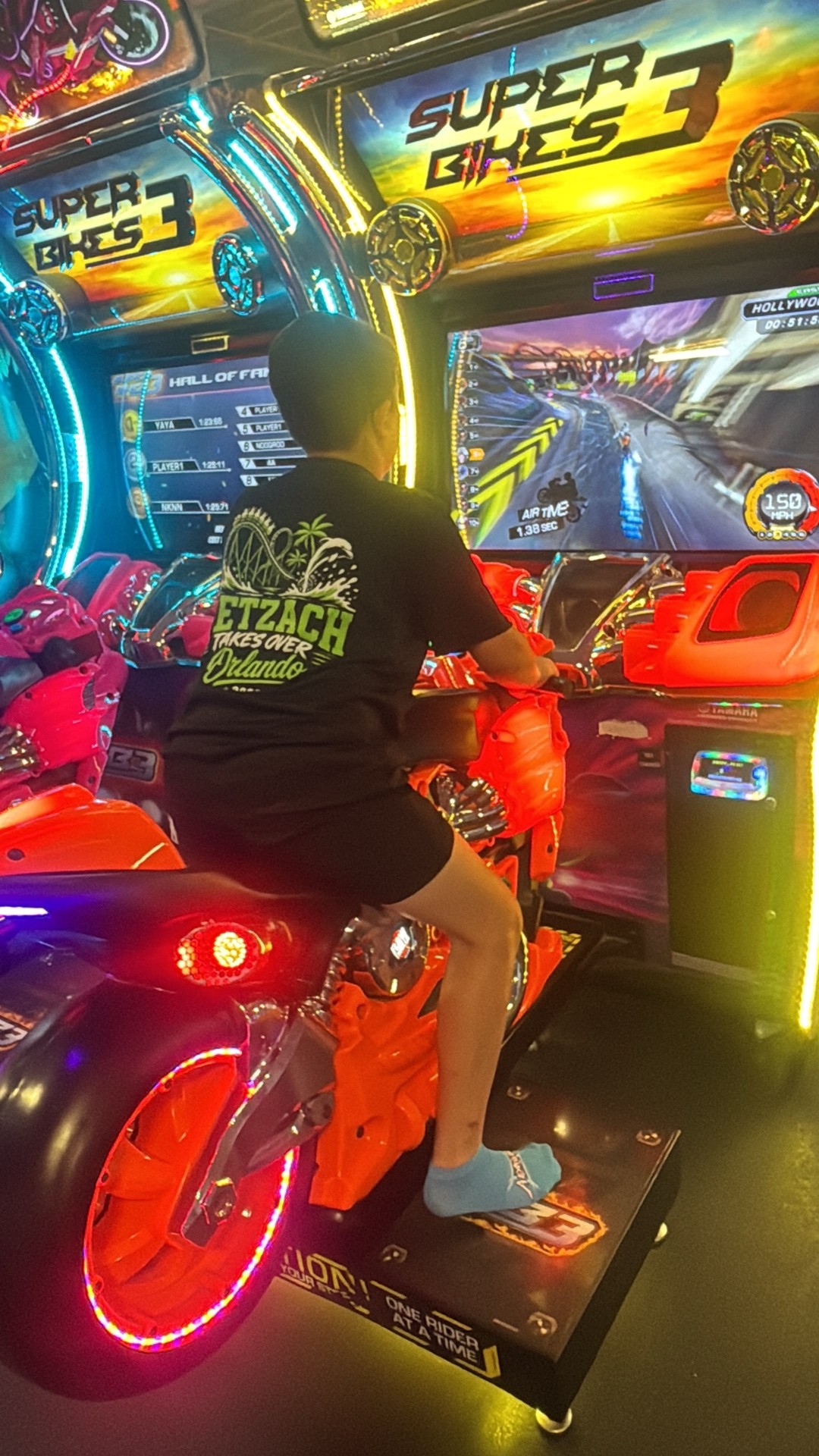 A camper riding an arcade motorcycle racing game in a dimly lit arcade