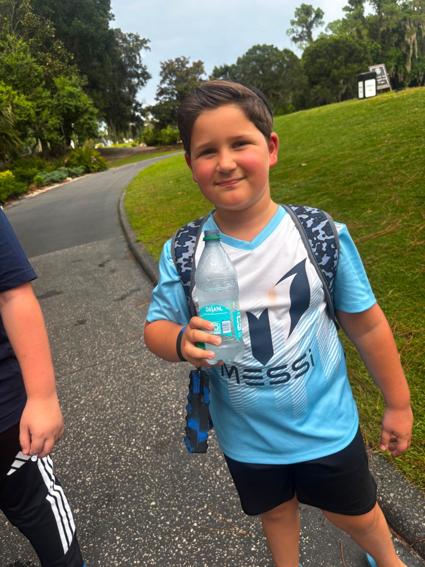A camper in a soccer jersey smiling with a water bottle on a golf course path
