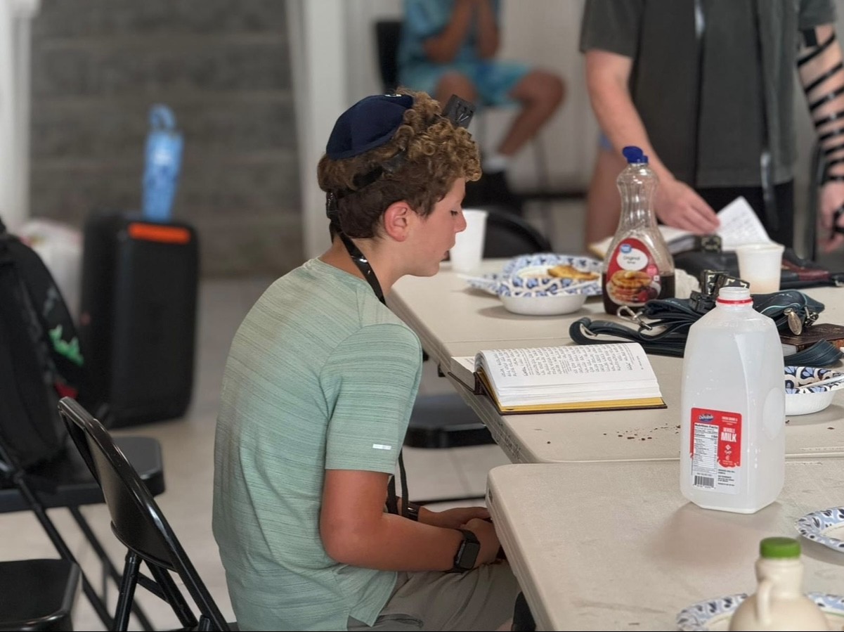 A camper wearing tefillin reading from a prayer book at breakfast